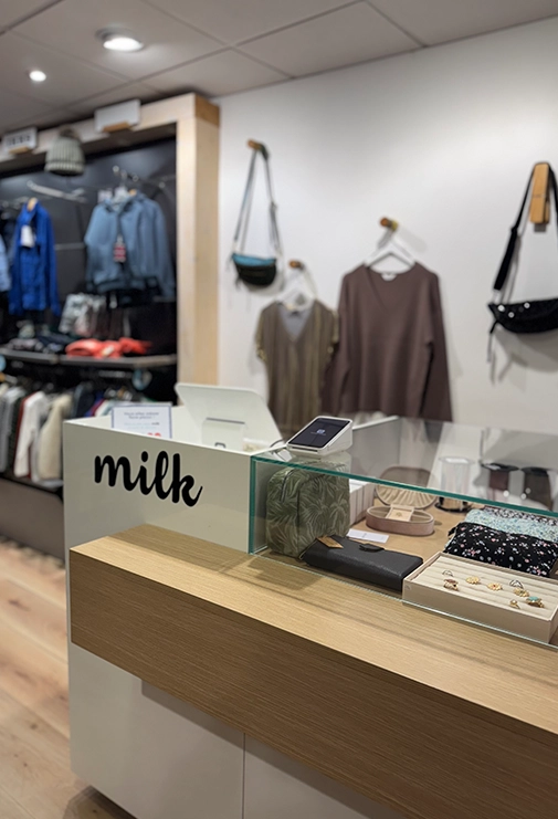 Dans l'image, on peut voir une boutique appelée 'Milk'. Au premier plan, il y a une table sur laquelle repose un chapeau rose. Ce chapeau attire immédiatement l'attention avec sa couleur vive et sa forme élégante. À l'arrière-plan, on peut voir une penderie remplie de vêtements soigneusement disposés. Les vêtements sont suspendus à des cintres et sont variés en couleur et en style. L'ensemble de l'image dégage une ambiance chic et tendance, invitant les clients à découvrir les articles proposés dans cette boutique.